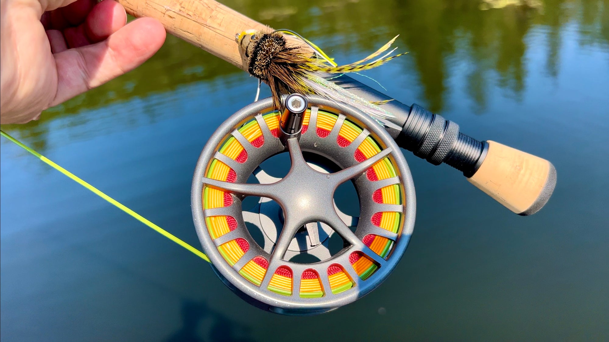 A hand holding up the bottom of a fly rod. The fly line is neon green, yellow, and red. There is water in the background.