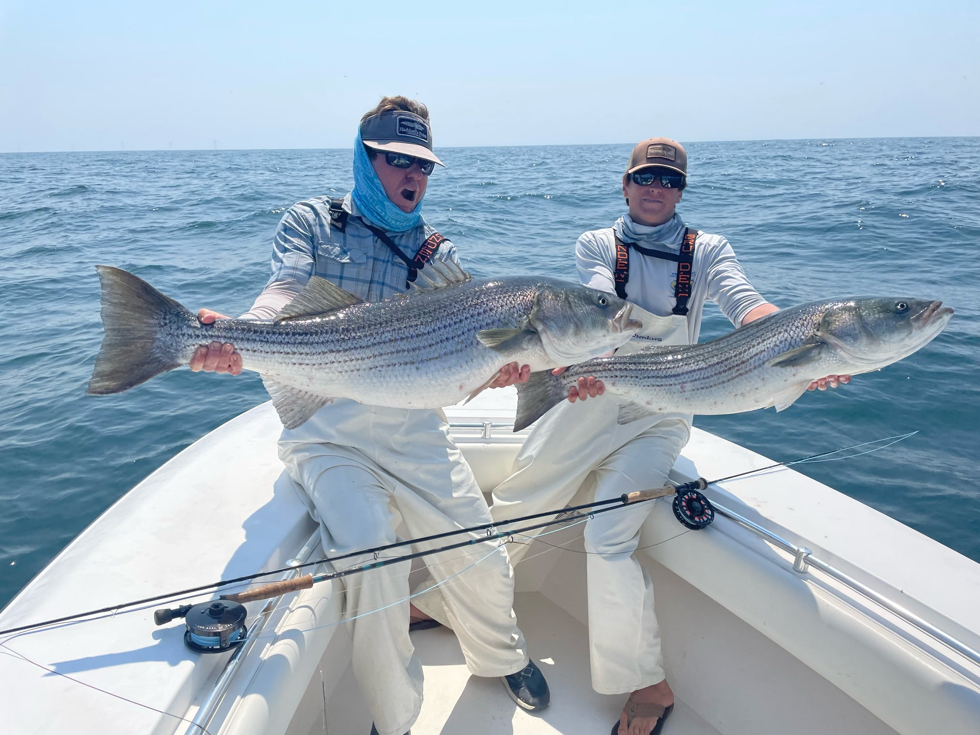 Two anglers holding up two large striped bass that they caught, they are on a boat with blue water in the background