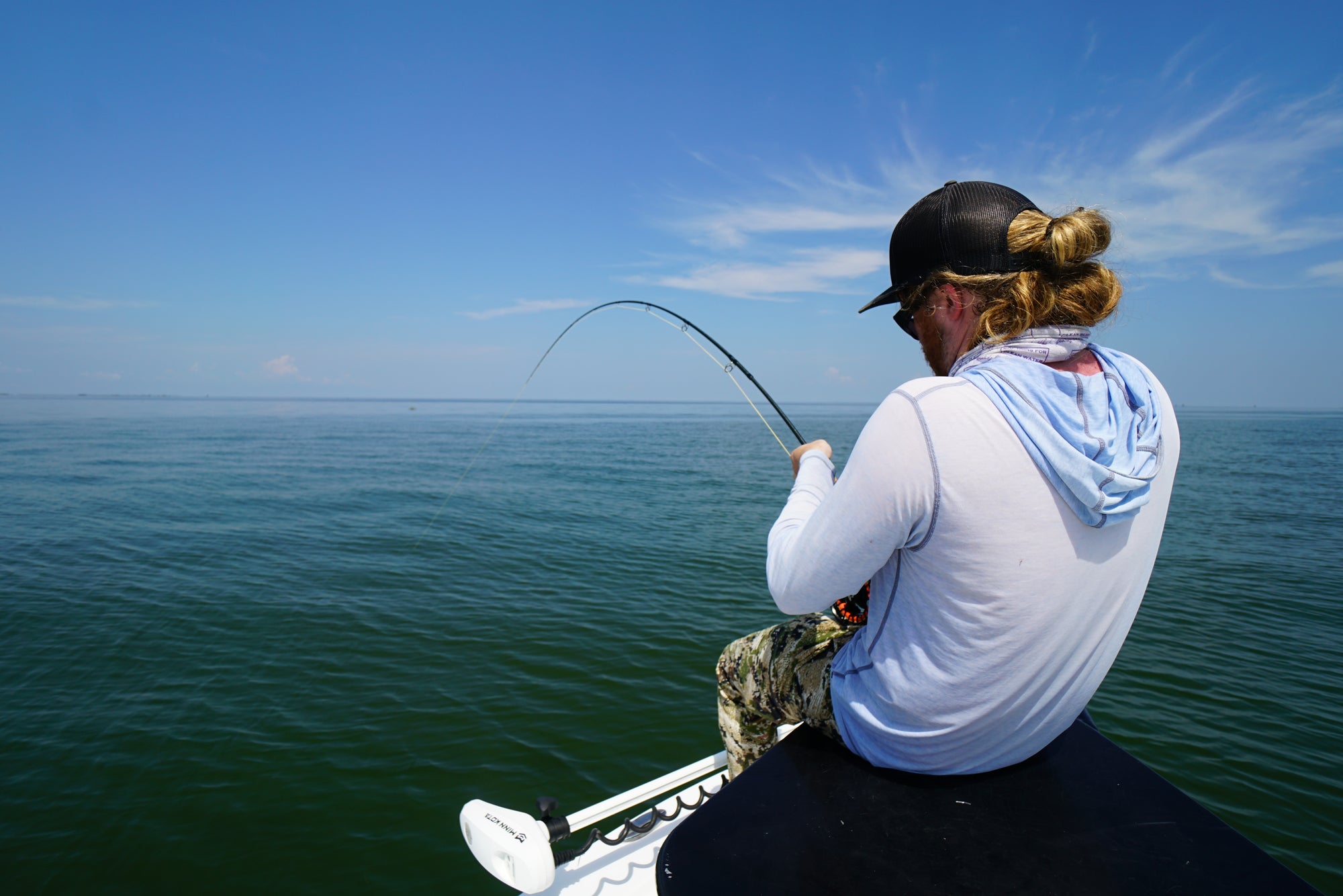 An angler is seated on a boat with his back towards the camera. He is reeling in his line.