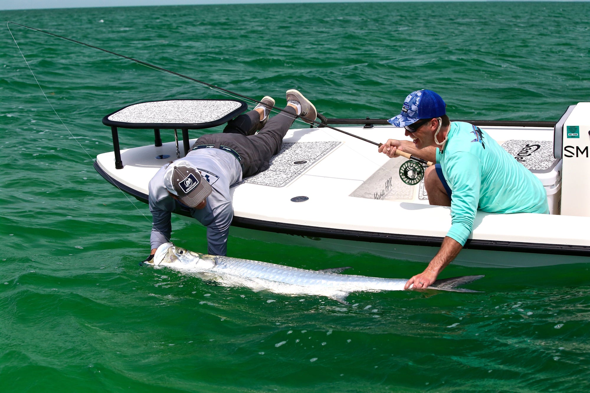 Two individuals are leaning over a boat and holding onto a tarpon.