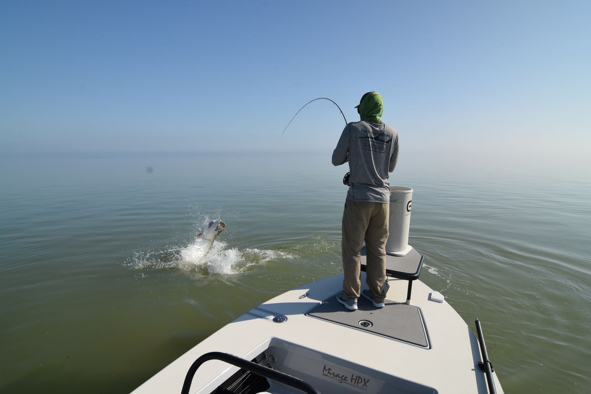 An angler is reeling in his fishing line. Meanwhile, the tarpon is jumping out of the water.