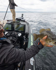There is an angler sitting on a boat and holding up a fish. And another individual fishing at the bow of the boat. 