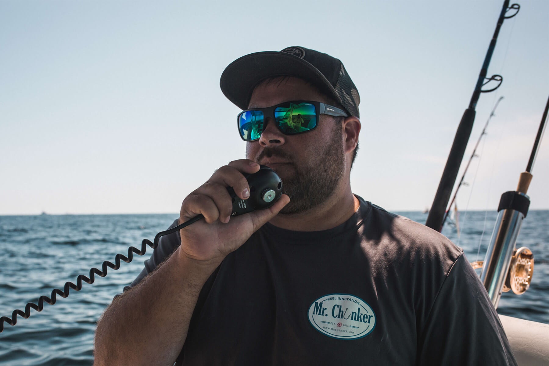 Jeff Warford is holding up a radio up to his mouth. There are two fishing rods behind him and water. 